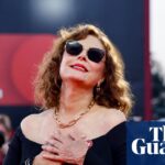 Susan Sarandon poses in sunglasses and an off-shoulder black dress as photographers shoot behind her