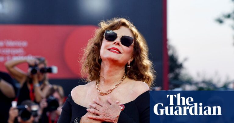 Susan Sarandon poses in sunglasses and an off-shoulder black dress as photographers shoot behind her